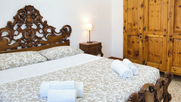 Een net opgemaakt bed met een bedsprei met dessin, gerolde handdoeken, een houten nachtkastje met een lamp en een grote houten garderobe.