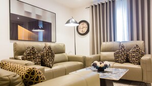 A modern living room with two beige sofas, patterned throw pillows, a coffee table with decorative items, a floor lamp, a wall clock, and a large abstract painting. - Comfy One Bedroom Apartment @ The Bantree (Accra)