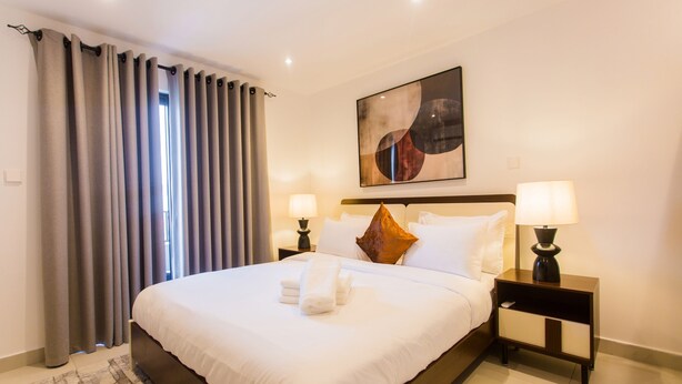 A hotel room with a large bed, two bedside tables with lamps, a painting on the wall, and a window with curtains. - Comfy One Bedroom Apartment @ The Bantree (Accra)