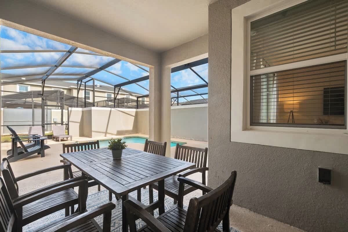 An outdoor patio area with a wooden table and chairs, a pool, and a covered seating area.