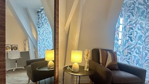 Un dormitorio tipo loft moderno con techo inclinado, un espejo grande, dos sillones, una mesita redonda con lámparas y una ventana con cortinas estampadas.