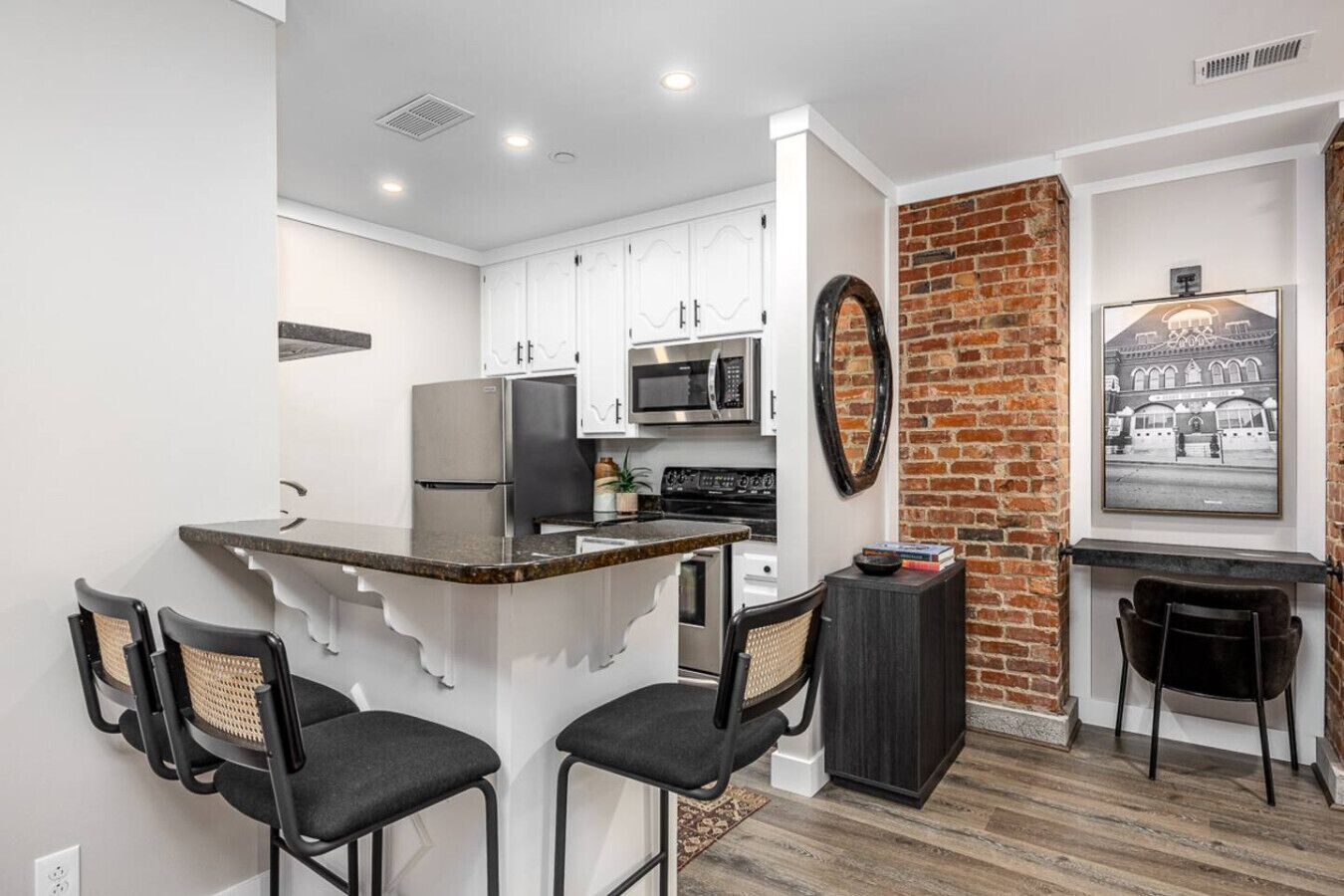 Une cuisine moderne avec des armoires blanches, un réfrigérateur noir, un four à micro-ondes et une cuisinière. Un mur en briques apparentes et un tableau encadré ornent le mur.