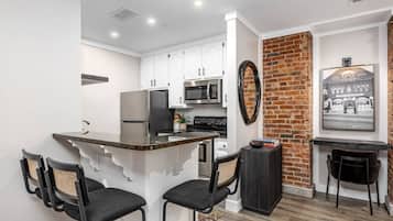Une cuisine moderne avec des armoires blanches, un réfrigérateur noir, un four à micro-ondes et une cuisinière. Un mur en briques apparentes et un tableau encadré ornent le mur.