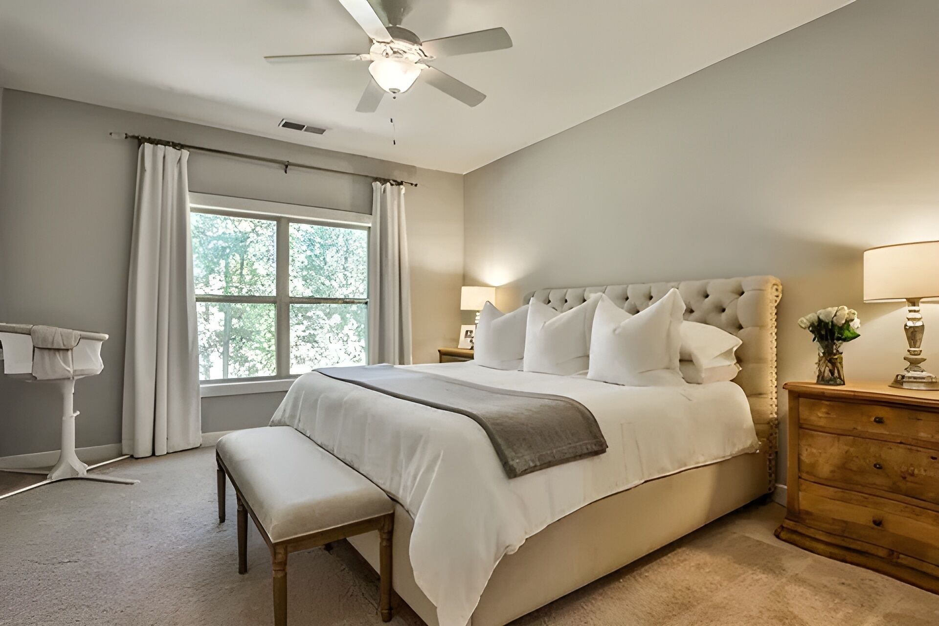 A bedroom with a large bed, a ceiling fan, a window with curtains, a bedside table with a lamp, and a bench.