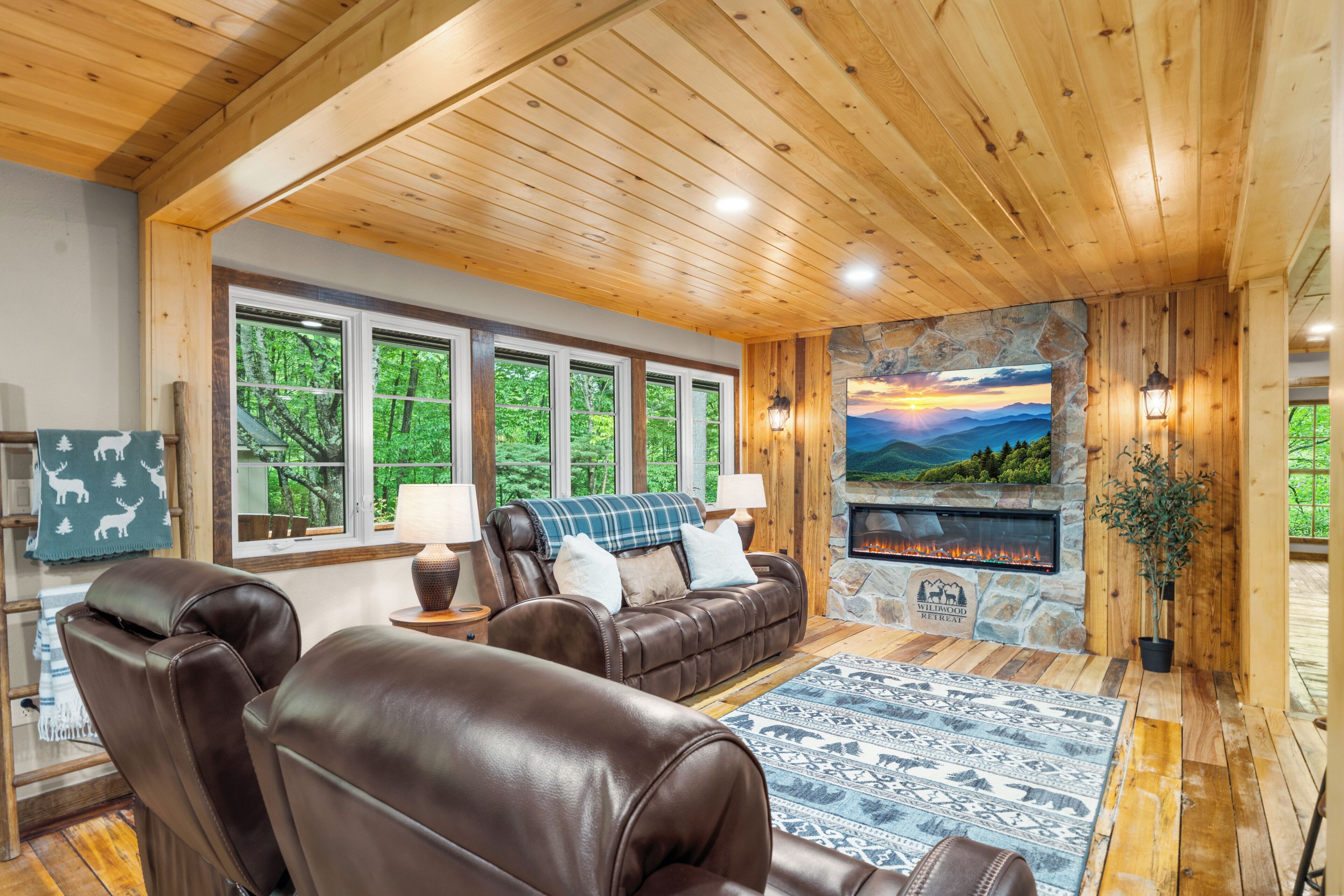 A cozy living room with a stone fireplace, leather sofas, and large windows offering a view of the forest.