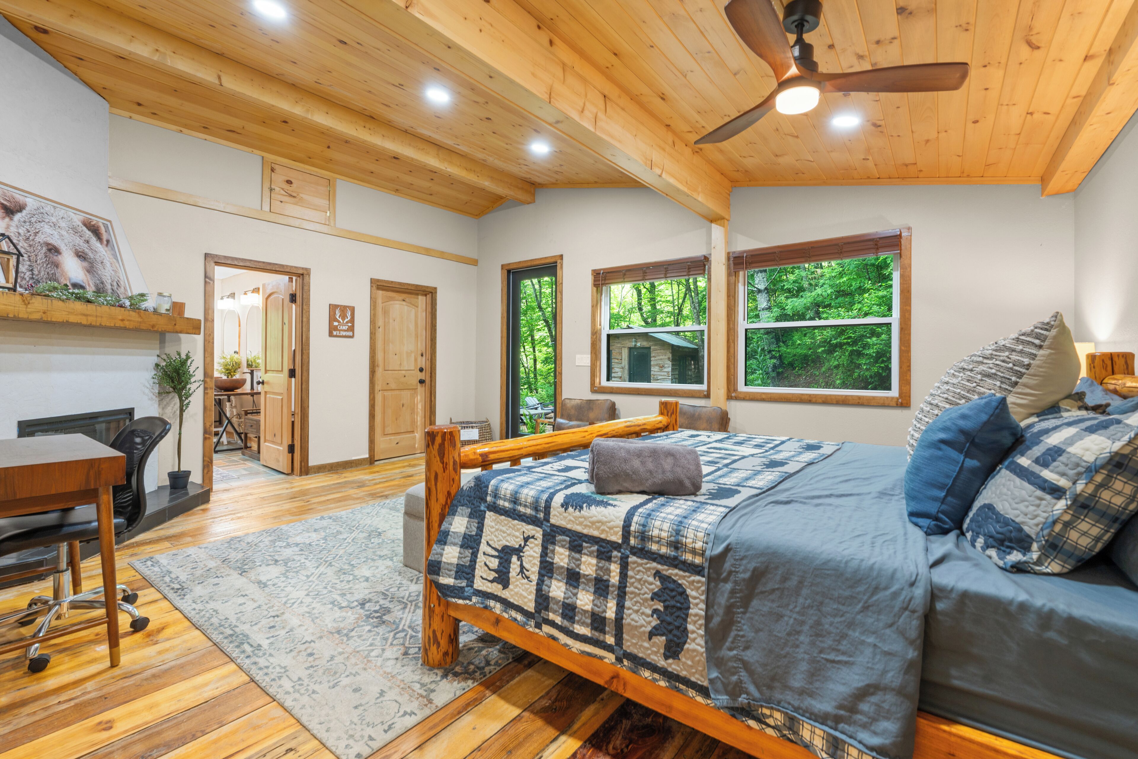 A bedroom with a wooden bed, a desk, and a chair. There are two windows with views of greenery.