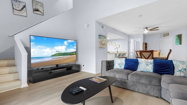 Ein modernes Wohnzimmer mit Flachbildfernseher, einem grauen Sofa mit gemusterten Kissen, einem schwarzen Couchtisch und einer Treppe, die in eine obere Ebene führt.