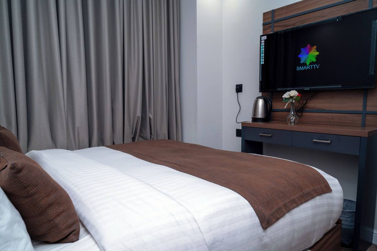 Une chambre d’hôtel avec un lit, un téléviseur fixé au mur, un bureau agrémenté d’un vase de fleurs et une fenêtre avec des rideaux.