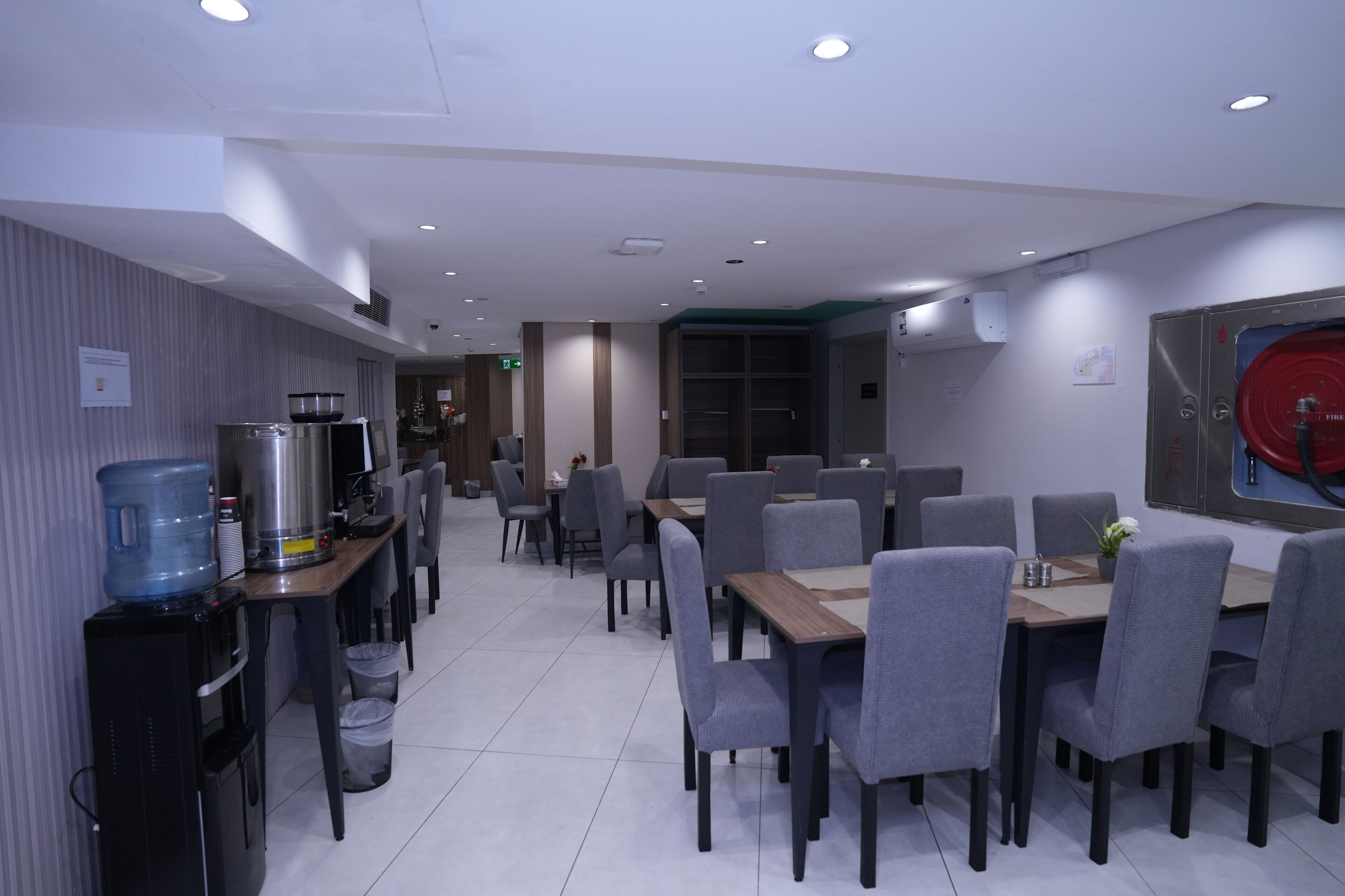 A modern dining area with grey chairs and tables, a water dispenser, and a fire extinguisher on the wall.