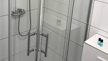 Eine gläserne Duschkabine mit Chromarmaturen, eine weiße geflieste Wand und ein kleines weißes Hängeschränkchen mit zwei Flaschen.