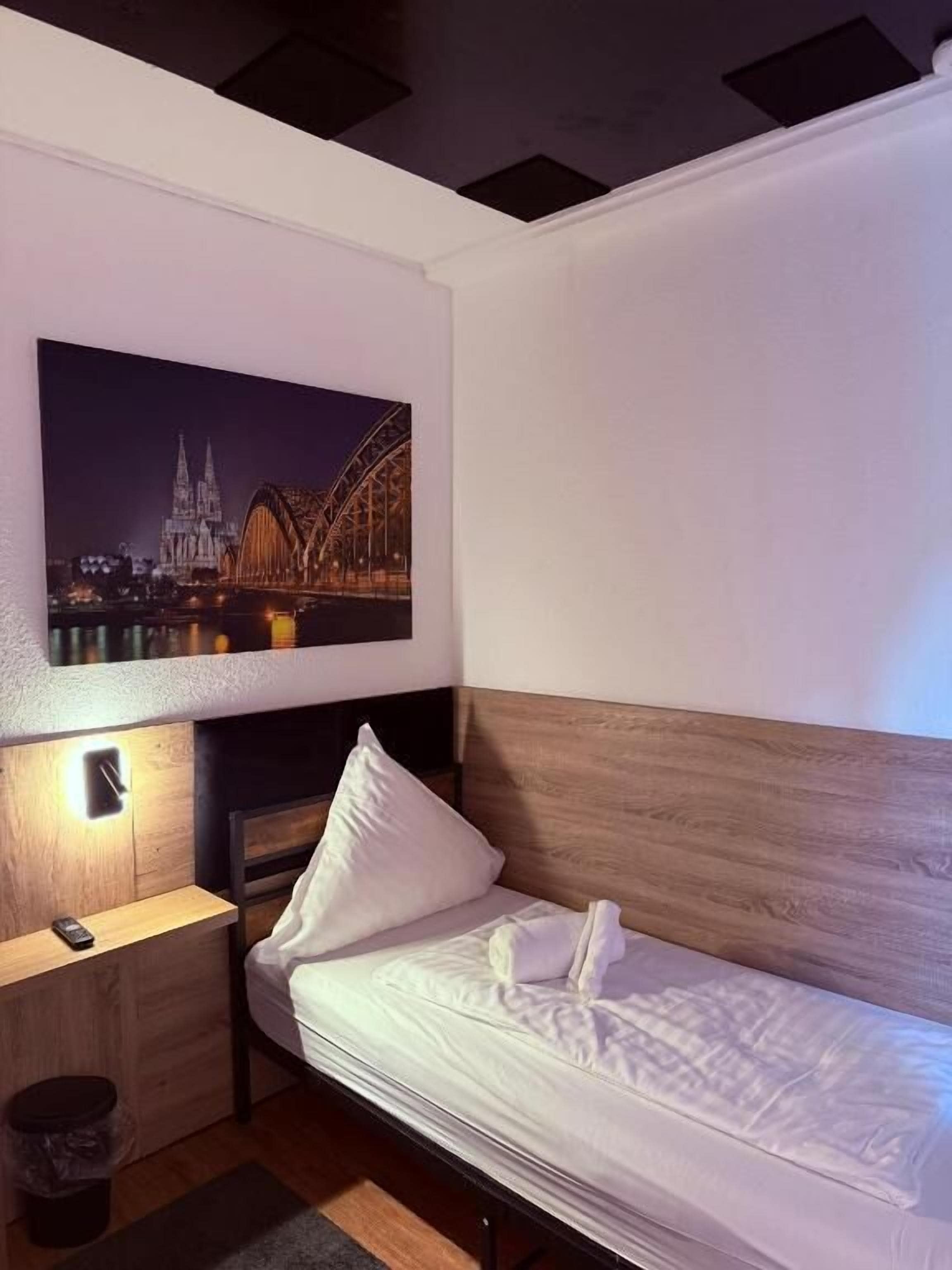 A single bed with a wooden headboard, a nightstand with a lamp, and a framed picture of a bridge at night.