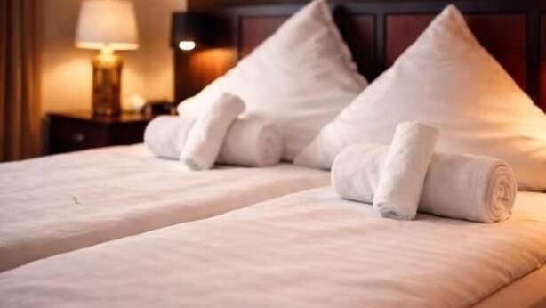 Ein Hotelzimmer mit zwei Betten, jedes mit einem ordentlich gefalteten weißen Handtuch, einer Lampe auf einem Nachttisch und einem dunklen Kopfteil.