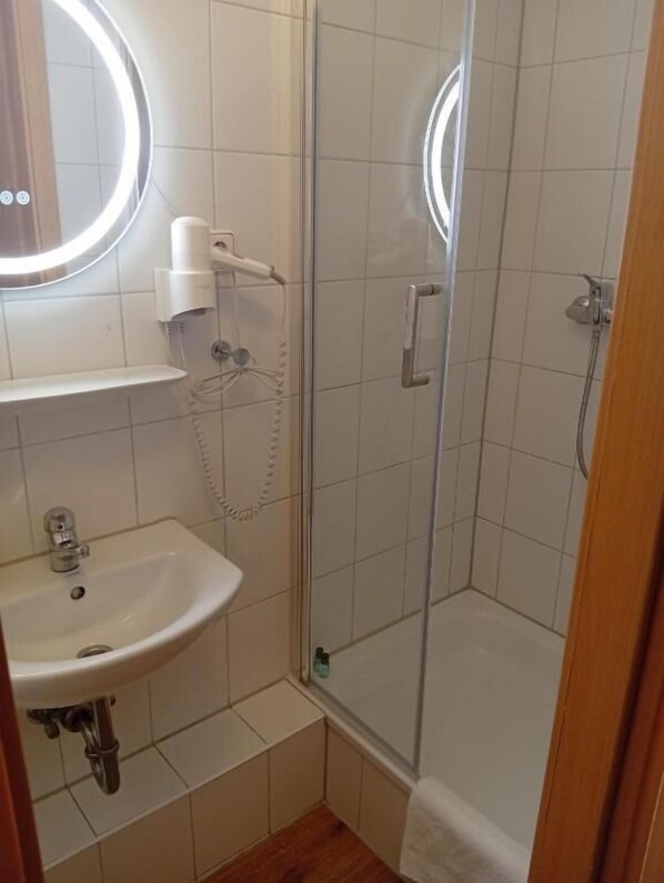 A bathroom with a glass-enclosed shower, a pedestal sink, and a mirror with a circular light.