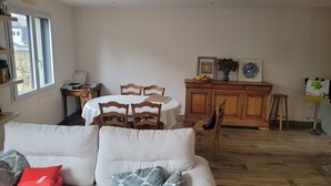 A living room with a white sofa, wooden dining table, and chairs. There is a wooden cabinet with a vase and framed pictures. A window provides natural light. - Villa near town center (approx. 600 m) with land (Dinan)