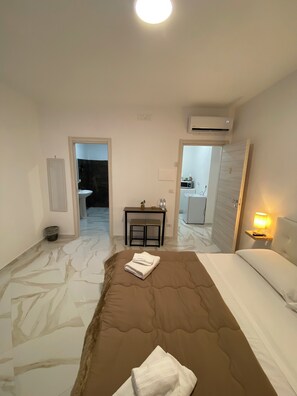 A hotel room with a bed, a bedside table, a lamp, and an air conditioning unit.