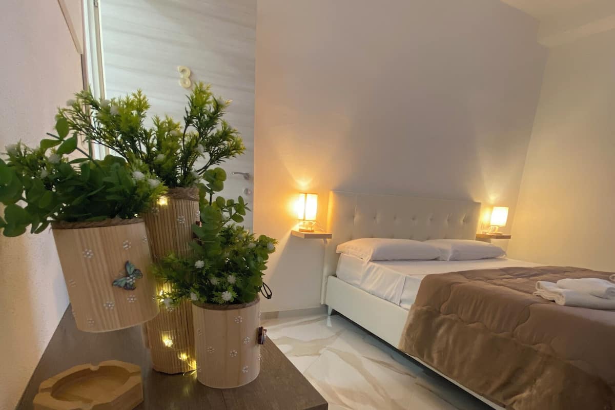 A hotel room with a bed, bedside tables, a desk with a plant, and a wall-mounted air conditioner.