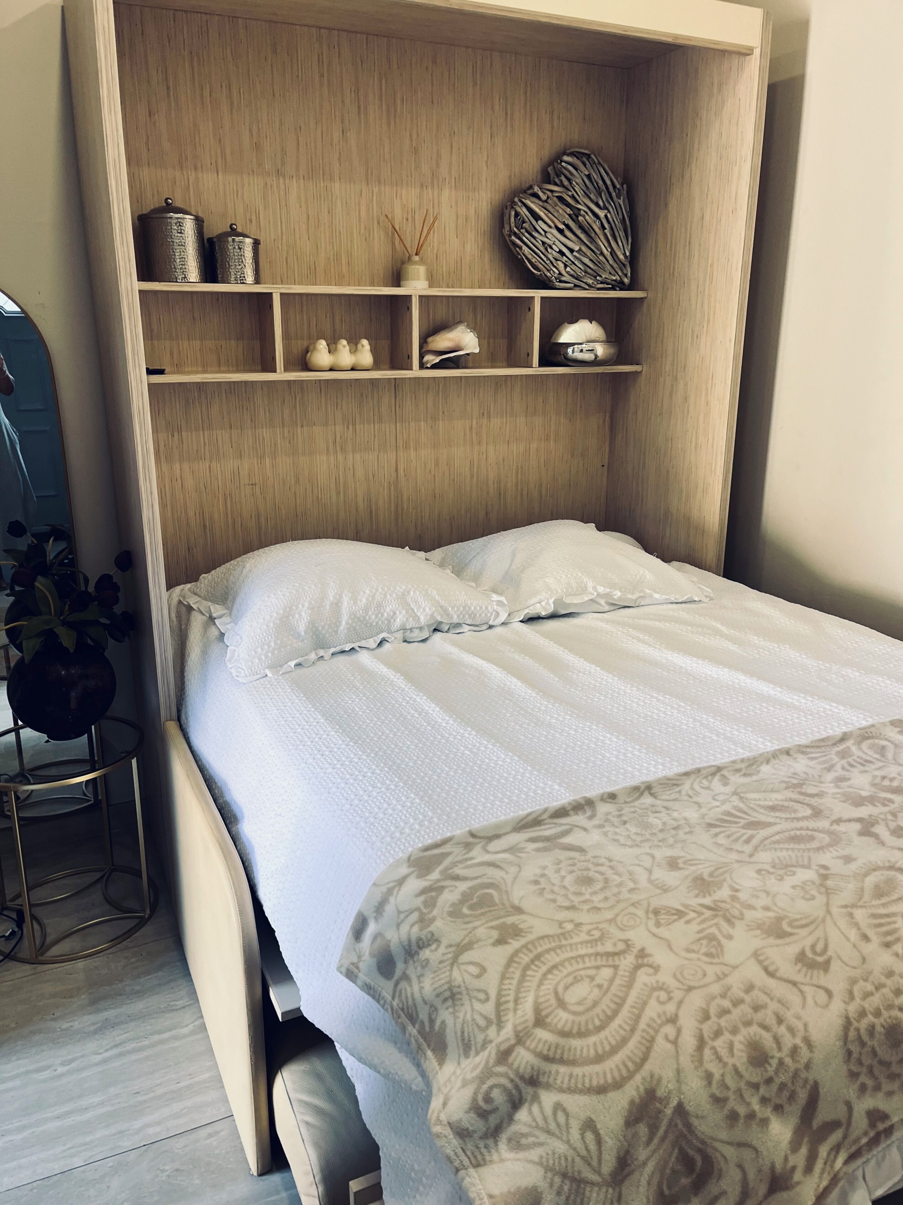 A bedroom with a bed, a wooden headboard, shelves with decorative items, and a small table with a plant.