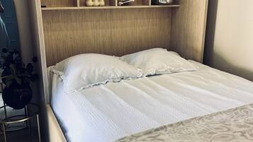 A bedroom with a bed, a wooden headboard, shelves with decorative items, and a small table with a plant.