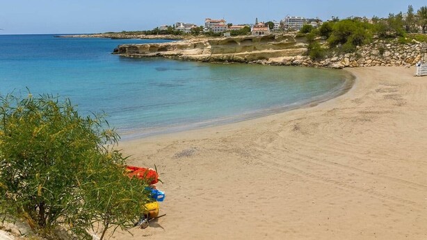 Ein sandiger Strand mit klarem, blauem Wasser, felsigen Klippen und Gebäuden im Hintergrund.