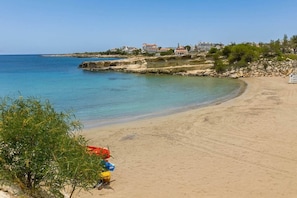 Ein sandiger Strand mit klarem, blauem Wasser, felsigen Klippen und Gebäuden im Hintergrund.