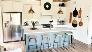 Une cuisine moderne dotée d’un réfrigérateur en acier inoxydable, d’un îlot central avec des tabourets, de luminaires suspendus et de divers ustensiles de cuisine accrochés aux murs.