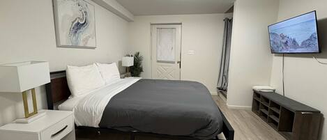 A modern bedroom with a bed, bedside table, lamp, TV, and a door leading to another room.
