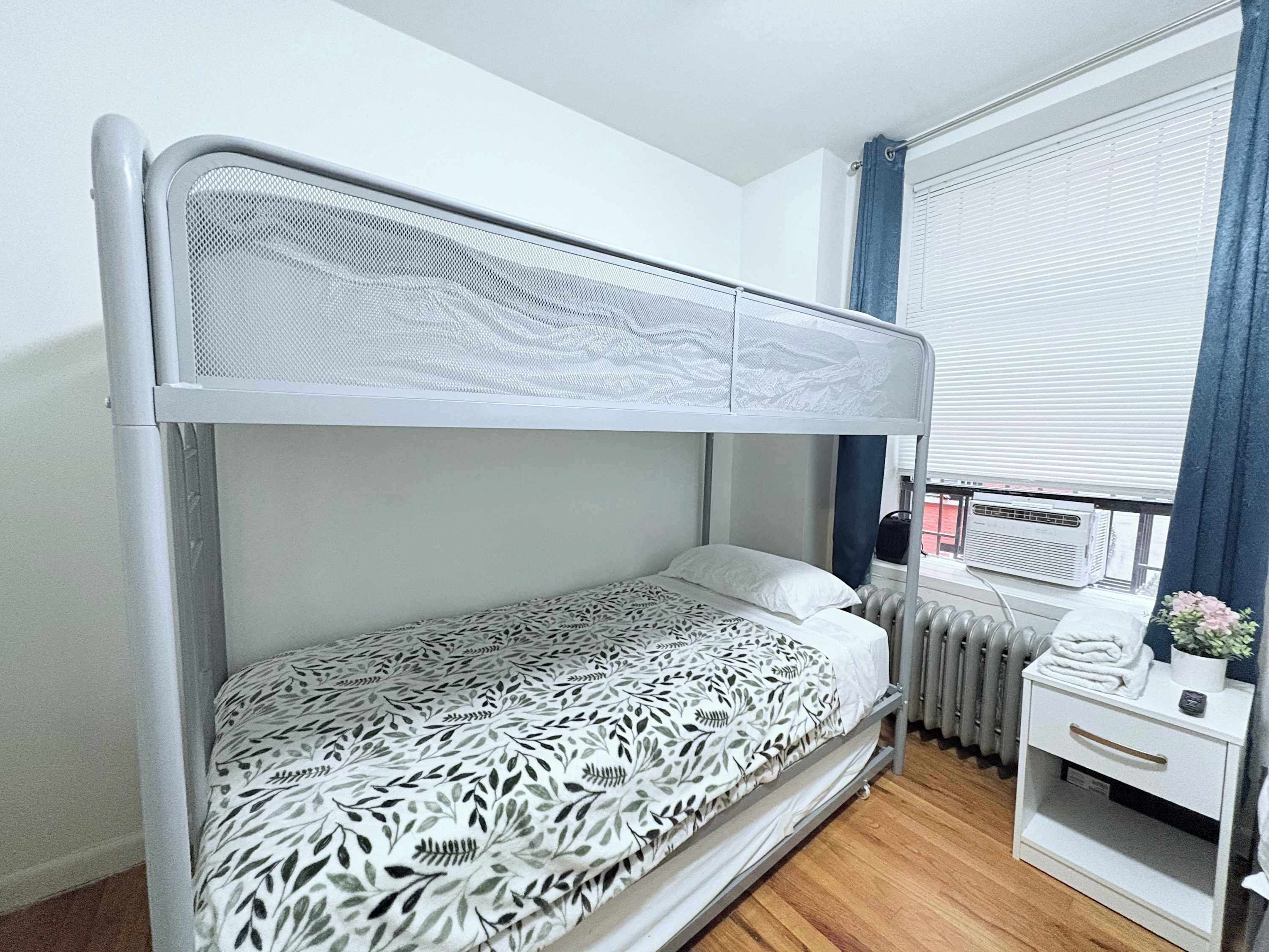 A bunk bed with a patterned bedspread, a white nightstand with a drawer, and a window with blinds.