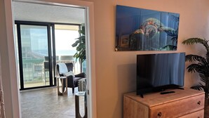 Ein Wohnzimmer mit Fernseher auf einer Holzkommode, ein Balkon mit Stühlen und einer Pflanze sowie ein Gemälde an der Wand.