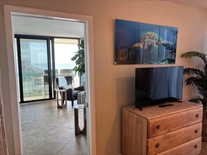 Una sala de estar con un televisor sobre un mueble de madera, un balcón con sillas y una planta, y un cuadro en la pared.