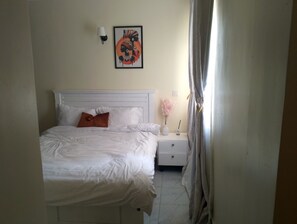 Una cama bien hecha con sábanas blancas y una única almohada marrón, una mesita de noche blanca con un florero de flores rosas, y una obra enmarcada sobre la cama.