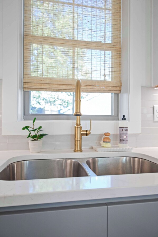 A kitchen with a stainless steel sink, a gold faucet, a window with blinds, a potted plant, and a dish soap bottle.