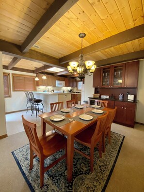Un coin repas avec une table en bois dressée pour six personnes, entourée de chaises, dans une pièce dotée de poutres apparentes en bois au plafond et d’un lustre.