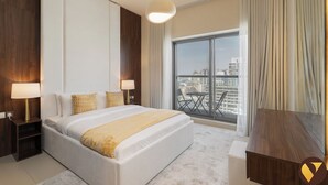 A modern hotel room with a large bed, two bedside tables with lamps, a view of the city through a large window, and a balcony with a table and chairs. - Vogue | Jeewar By Badr JVC near Circle Mall (Dubai)