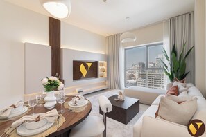 A modern hotel room with a dining area, a sofa, a large window with a city view, and a mounted artwork on the wall. - Vogue | Jeewar By Badr JVC near Circle Mall (Dubai)