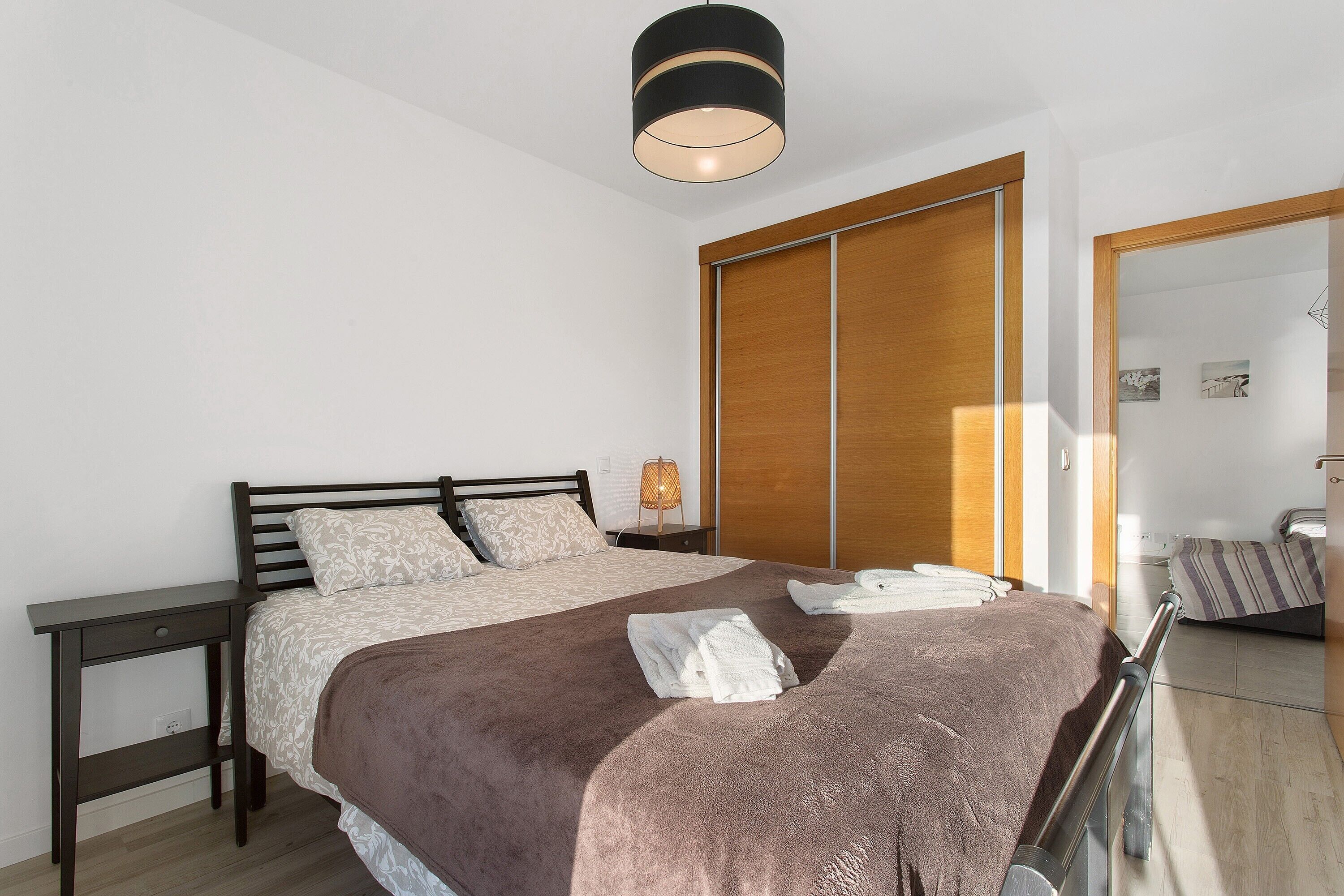 Een slaapkamer met een bed, nachtkastje, lamp, garderobe en uitzicht op een andere kamer.