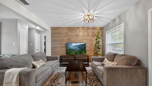 A living room with two grey sofas, a wooden coffee table, and a large TV screen. A Christmas tree is visible in the background. - 3-Bedroom Home Across from Busch Gardens (tampa)