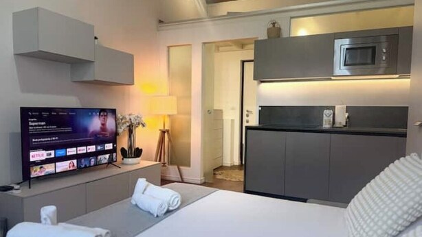 A hotel room with a bed, a flat-screen TV mounted on the wall, a microwave above the kitchen counter, and a small table with a lamp and a potted plant. - A Casa di Imma - Elegant studio apartment in Porta Rom (Milano)
