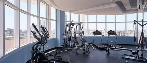 Ett gym med olika typer av träningsutrustning, stora fönster och utsikt över staden.