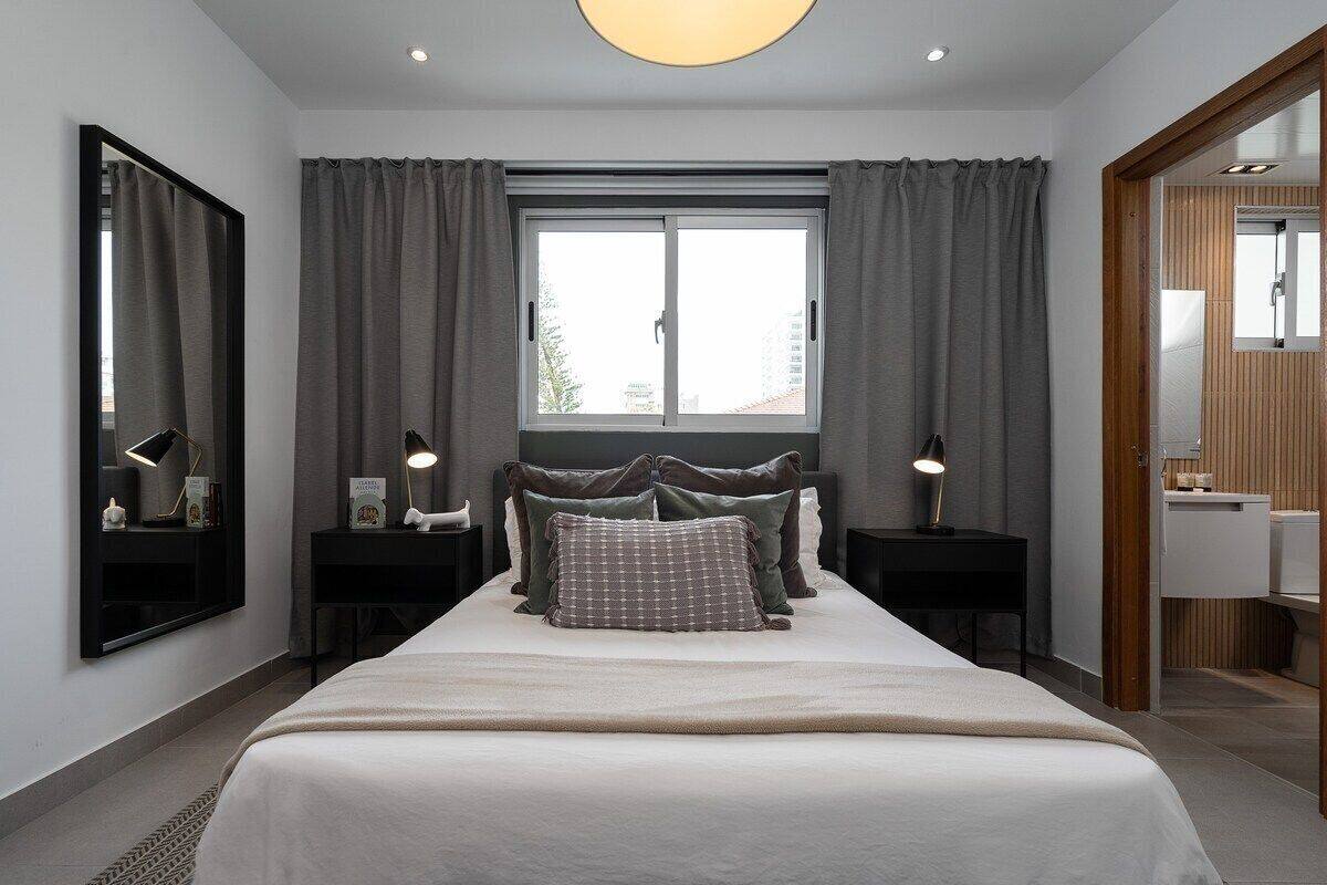 A modern bedroom with a large bed, two bedside tables, a nightstand, a window with curtains, and a mirror.
