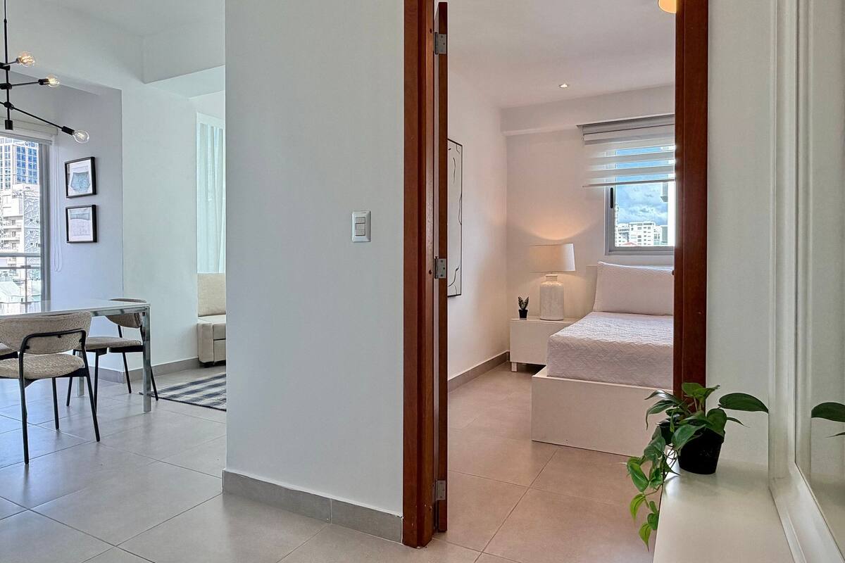 A modern bedroom with a bed, bedside table, lamp, and a view of the city through a window.