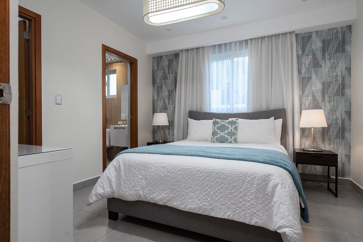 A modern hotel room with a large bed, two bedside tables with lamps, a mirror, and a view of the city through large windows.