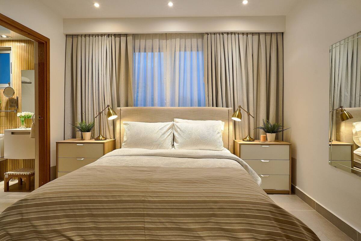 A neatly arranged bedroom with a large bed, two bedside tables with lamps, a mirror, and a window with curtains.