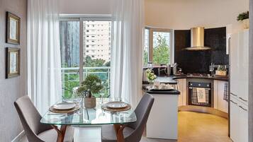 A modern kitchen with a glass dining table, two chairs, and a view of a cityscape through large windows.
