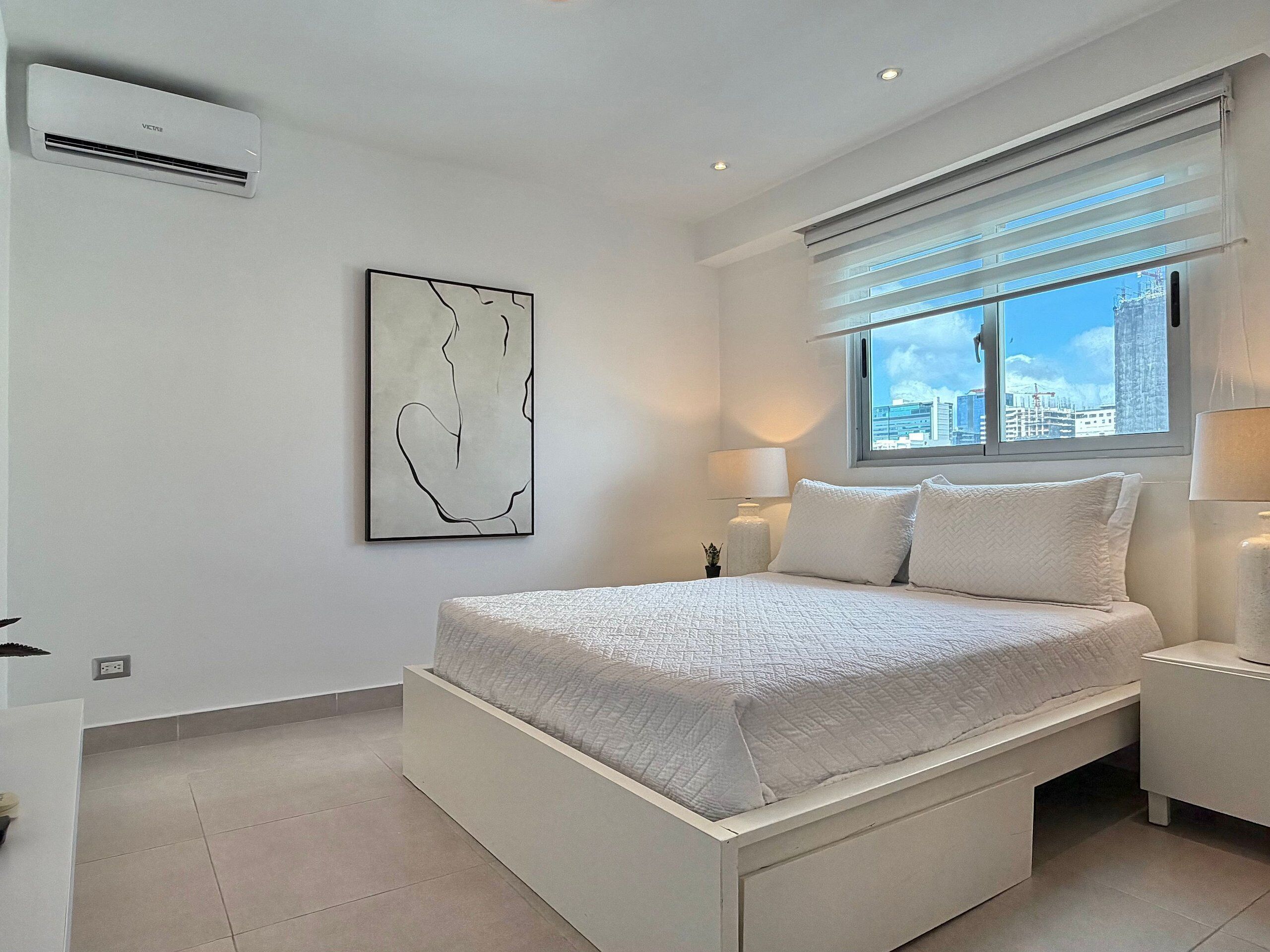 Un dormitorio moderno con cama, mesitas de noche, una ventana con persianas, un equipo de aire acondicionado y una obra de arte colgada en la pared.