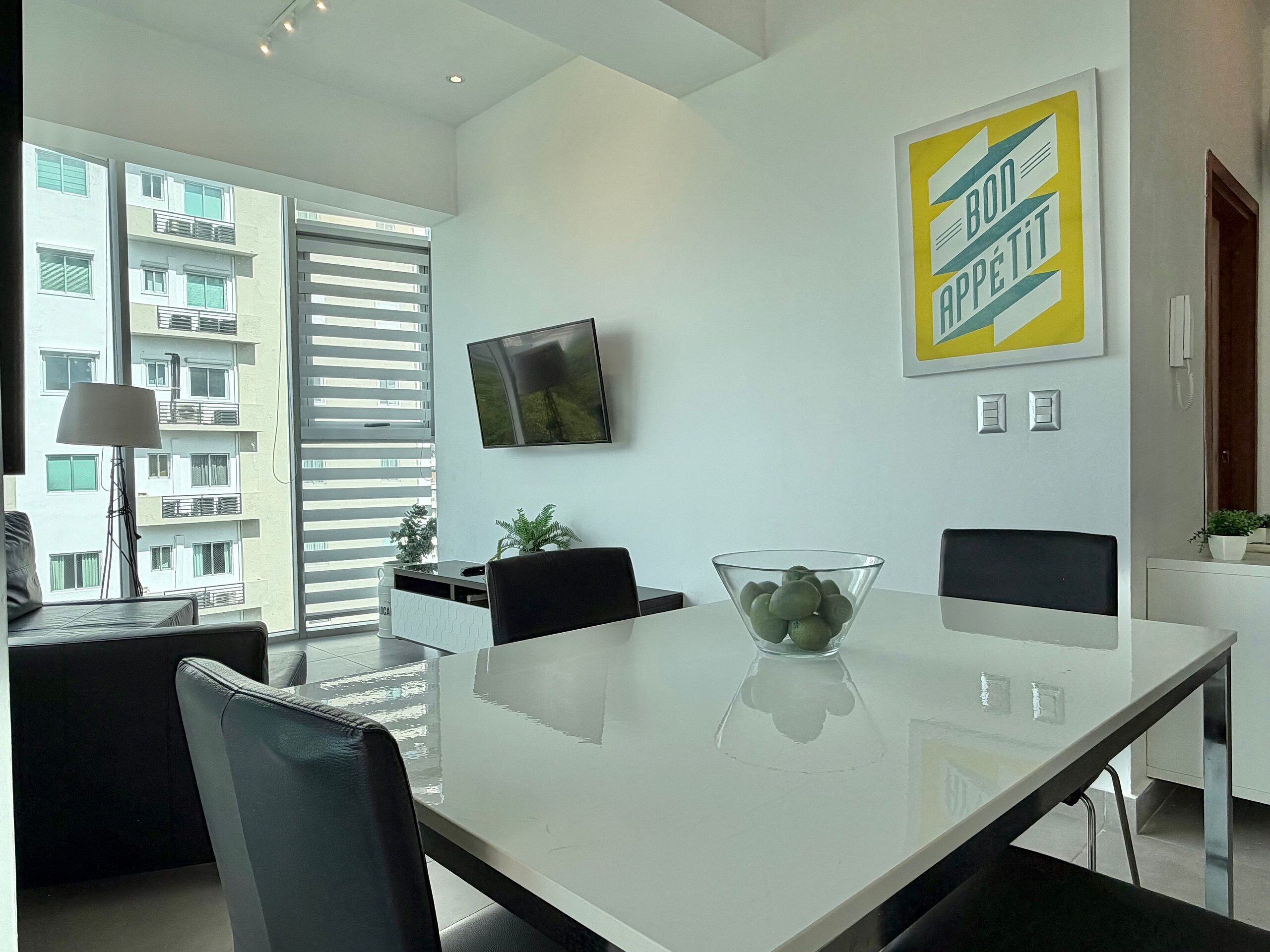 A modern dining area with a glass table, black chairs, and a bowl of green apples. A TV is mounted on the wall, and a yellow sign with text is visible.