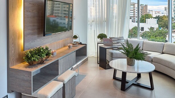 Una sala de estar moderna con un televisor grande montado en un panel de madera, un sofá, una mesa de centro y un balcón con vistas a la ciudad.