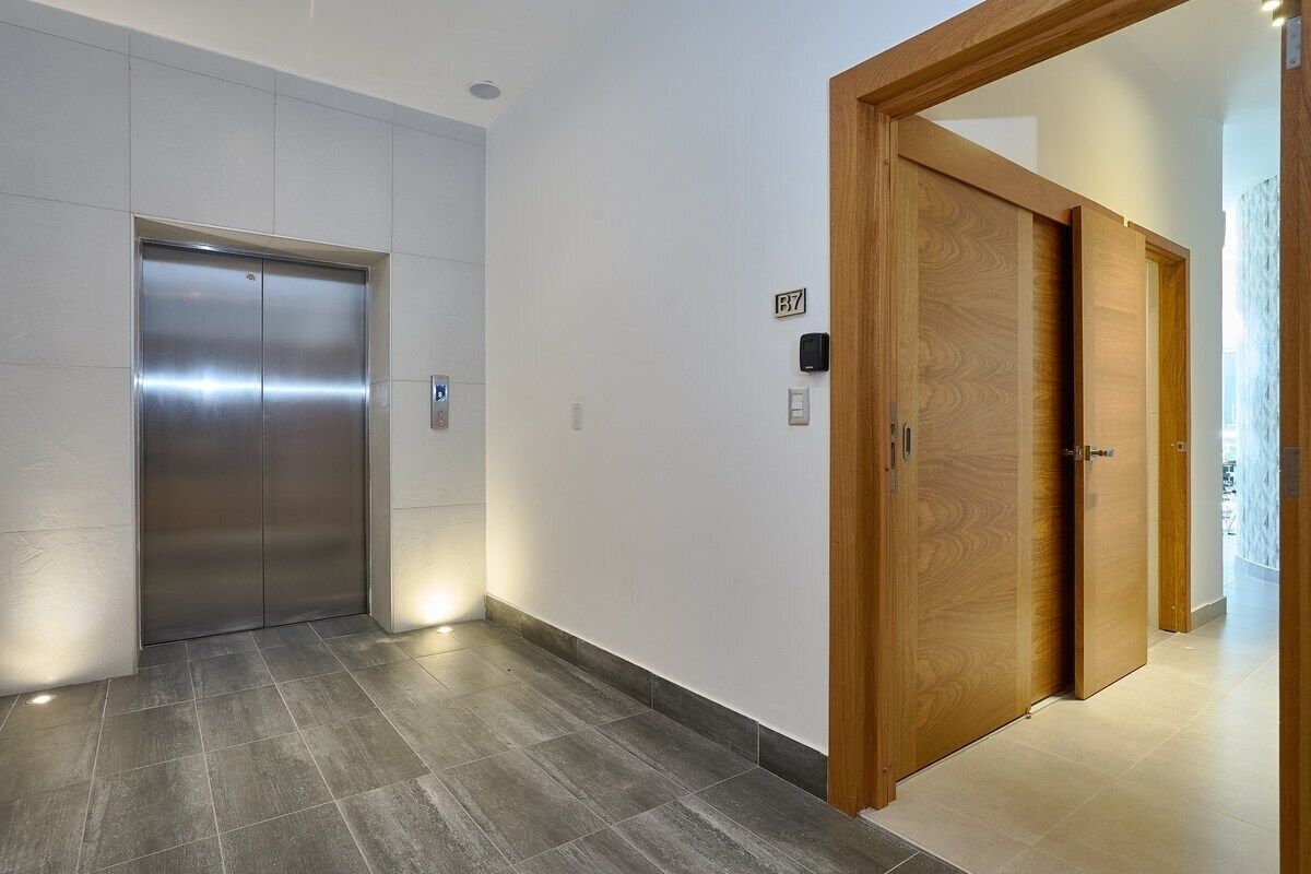 A hallway with a metallic elevator and wooden doors, a number sign 'B7', and a tiled floor.