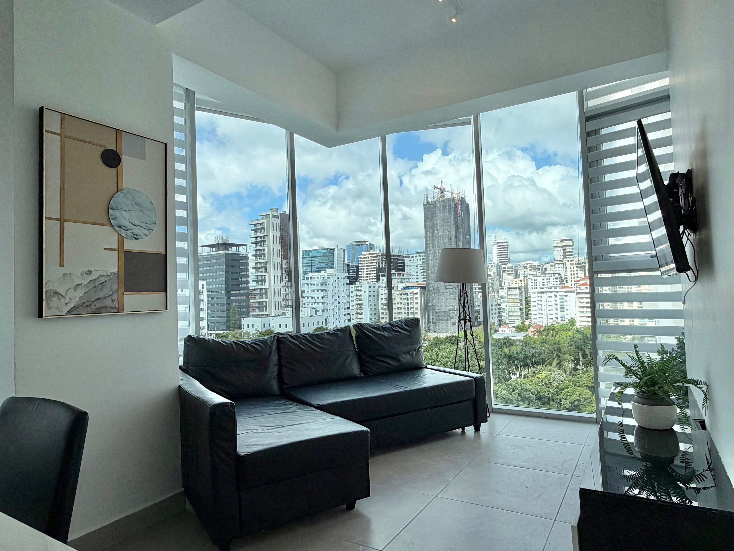 Una sala moderna con un sofá de cuero negro, una lámpara de pie y vistas a la ciudad a través de amplios ventanales.