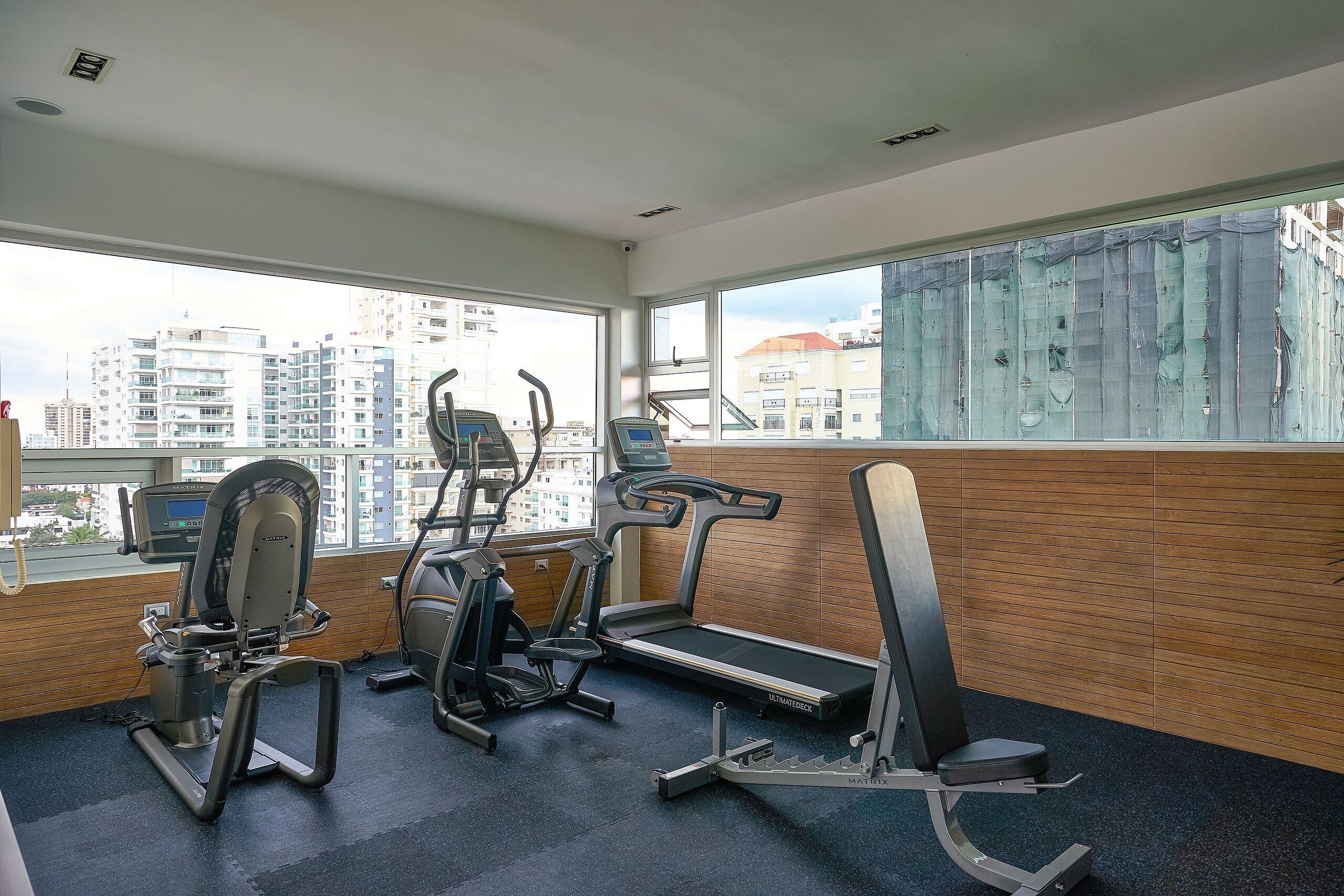A gym with treadmills, elliptical machines, and weight benches, featuring large windows with a city view.