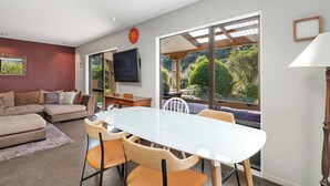 Una sala de estar moderna con un sofá, una mesa de comedor con sillas, un televisor y ventanas amplias que dan a vistas de follaje.
